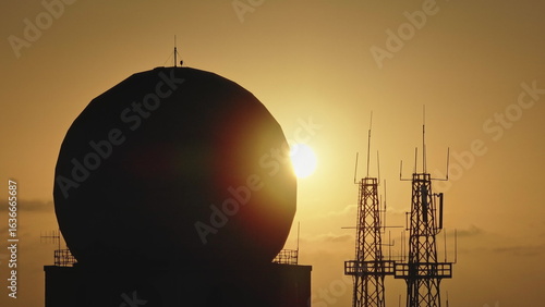 Malta: Silhouette of large radome and radio towers against a warm orange sunset sky in Malta, evoking a military or spy base atmosphere. Drone flight