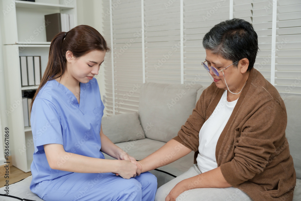 Fototapeta premium Young female therapist holding wrist of senior asian woman and examining forearm joint movement during physical therapy session while sitting on sofa in bright room