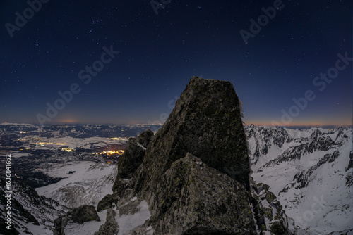 Aerial view of a craggy peak under a starlit sky, with the distant town lights twinkling like scattered jewels, Koncista, PreÅ¡ov Region, Slovakia.