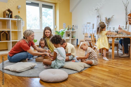 Two preschool teachers with children sitting on floor, engaging in collaborative early learning activity in bright classroom