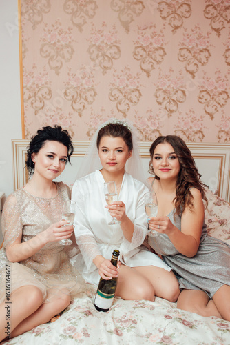 Bride in veil with bridesmaids sitting on the bed, raising glasses of champagne. Joyful, festive and friendly wedding morning atmosphere.