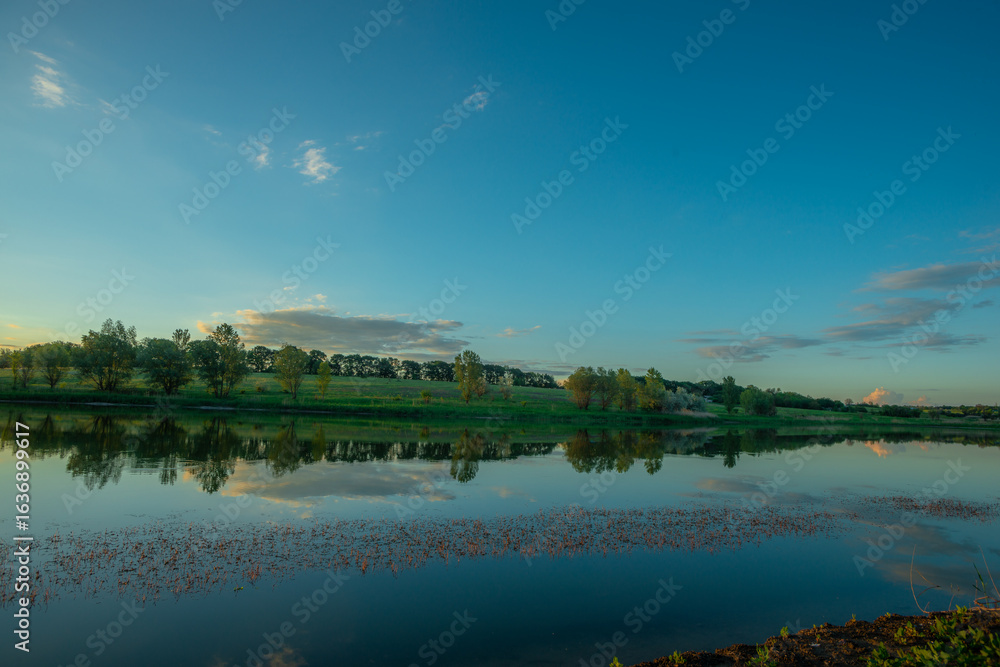 Obraz premium A wide horizontal view of a serene lake in the evening, with a clear blue sky and clouds reflected on the water, and a distant green treeline on the opposite shore