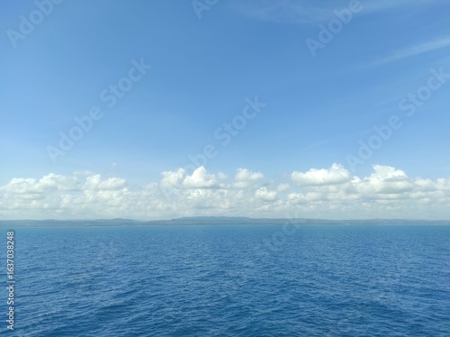 This scenic photograph presents a vast, open-water landscape under a clear blue sky. The deep blue ocean, marked by gentle ripples, stretches out to a distant horizon. 