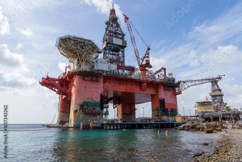 Fotografi Plataforma de petróleo offshore em Curaçao, vista de perto no porto, destacando estrutura industrial, guindastes e operação marítima sob céu parcialmente nublado