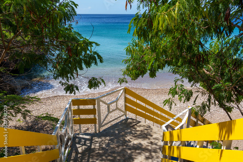 Escada com corrimões amarelos levando à praia de águas azul-turquesa em Curaçao, cercada por vegetação tropical e cenário paradisíaco do Caribe.