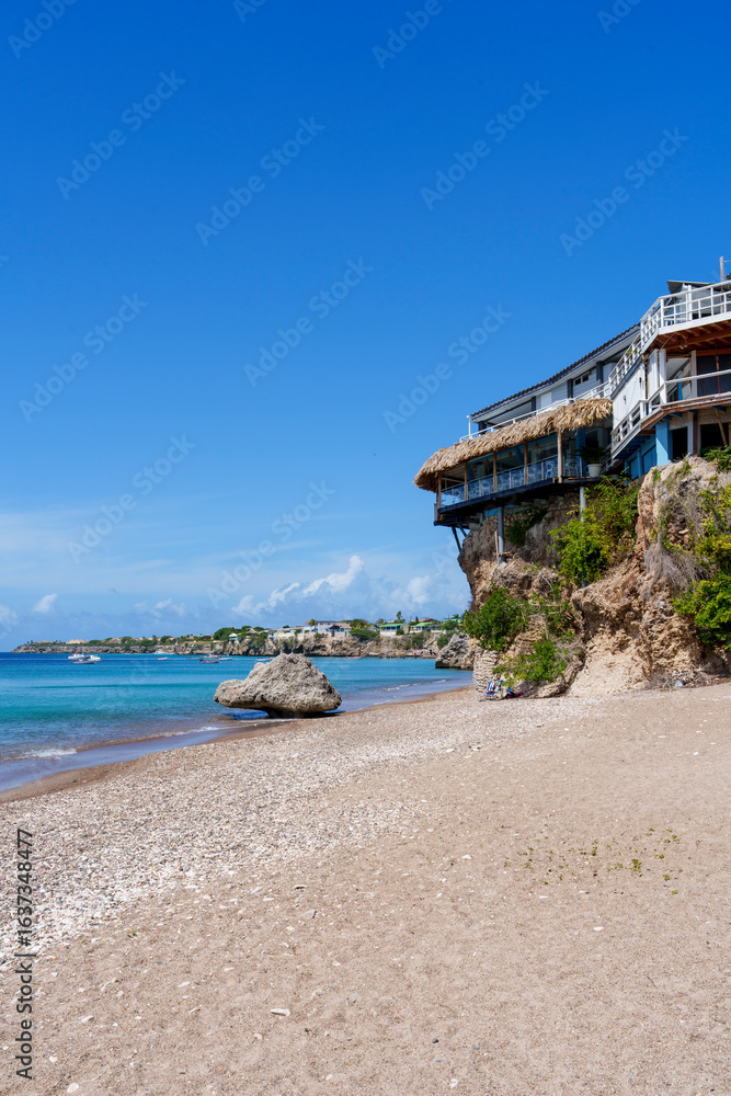Fototapeta premium Praia tropical em Curaçao com águas azul-turquesa, areia branca e turistas aproveitando o dia ensolarado, cercada por natureza e falésias rochosas.