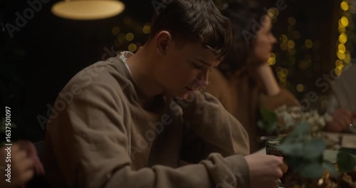 Sad teenage guy sitting at the table and looking at the ice while his little brother touches his shoulder during the family Christmas dinner in the evening