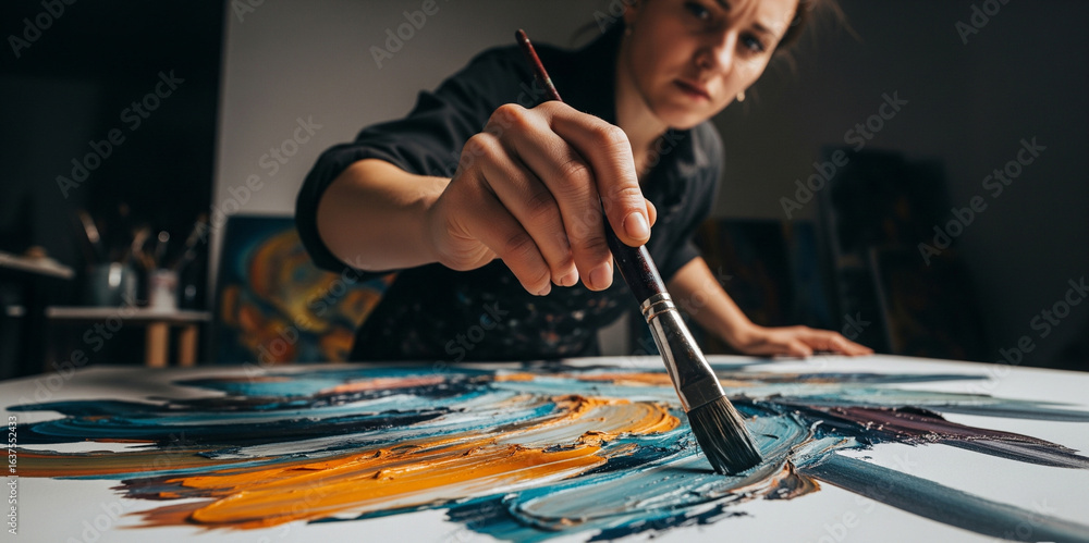 Fototapeta premium A female artist energetically applies bold yellow paint to a vibrant abstract oil painting in her dimly lit studio.