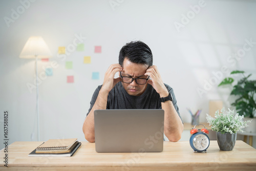 Slika na platnu Young man feeling stressed while working at home on a laptop, struggling to focus and under pressure in a bright and organized workspace