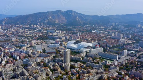 4K drone panorama of Marseille : the Orange Velodrome gleams amid dense city blocks, framed by Provencal mountains and Mediterranean light - epic urban skyline and sports landmark.