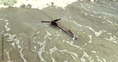 In slow motion, foamy water drifts over a rusty grapnel anchor, tied with a thick rope and submerged in the shallows of a sandy beach.