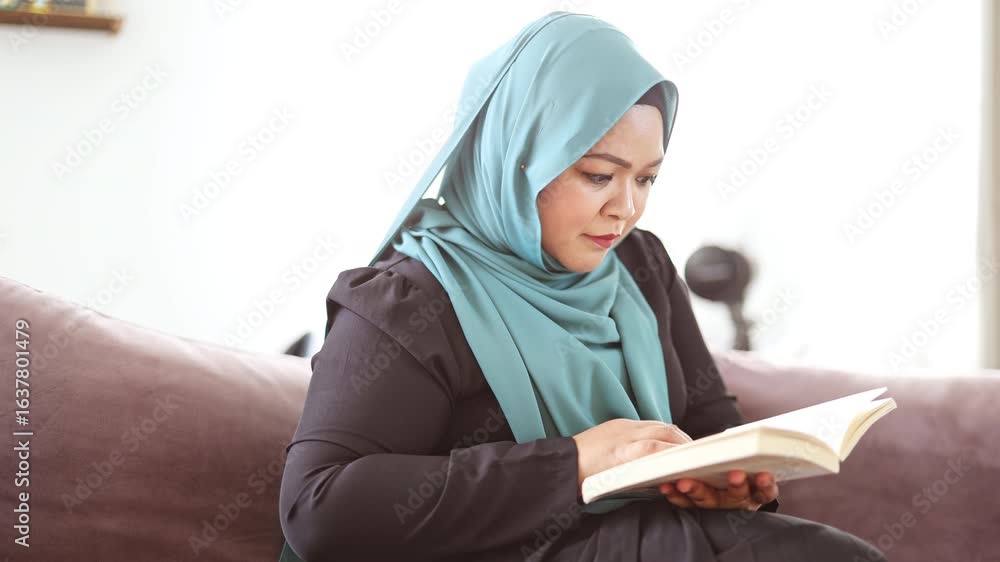 Woman in hijab reading a book while sitting on a sofa in her home

