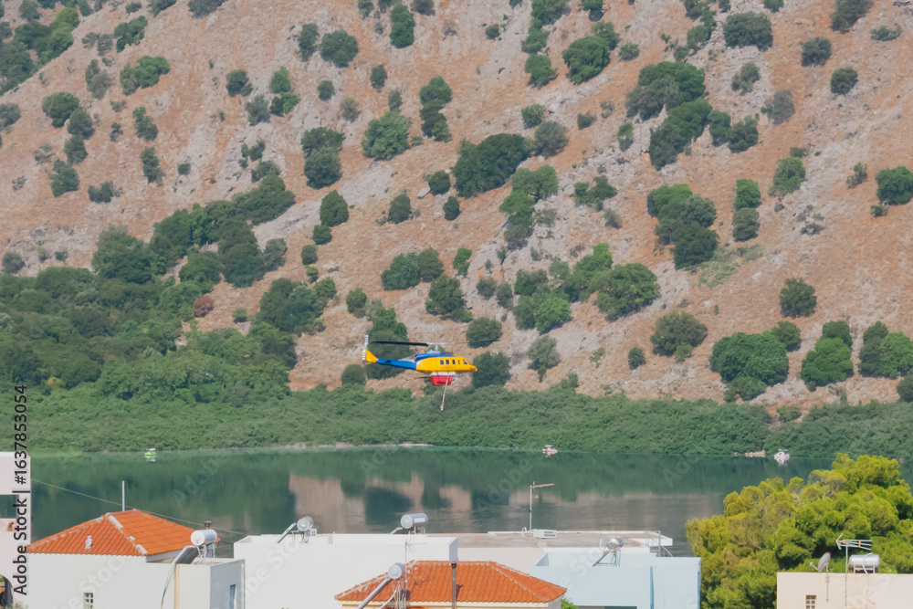 Fototapeta premium Blue and yellow firefighting helicopter flying over lake to collect water for extinguishing fire
