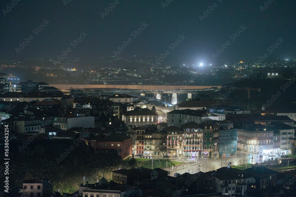 Obraz premium At night, buildings are illuminated along a narrow street. A bridge spans a wide river. Faint lights from distant hills twinkle. The sky above is dark, suggesting late hours