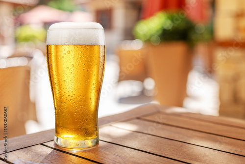 Obraz na plátne Glass of chilled beer on wooden table and blurred summer terrace at the background