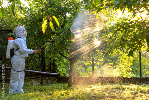 A gardener in a protective suit, mask and glasses uses a professional sprayer to treat the garden with insecticides and fungicides from pests