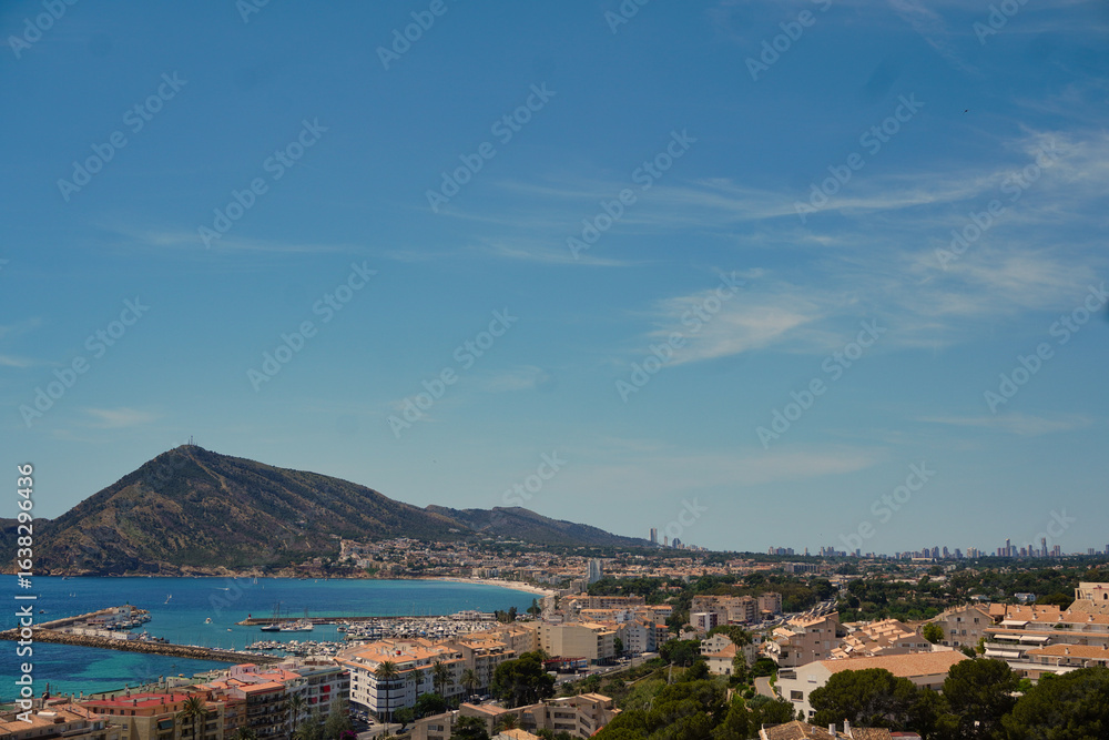 Obraz premium Panoramic view of the coast of Altea, Alicante, Spain, showing the marina, bay, and mountain landscape under a blue sky. Benidorm skyline is visible in the distant background on a sunny summer day.