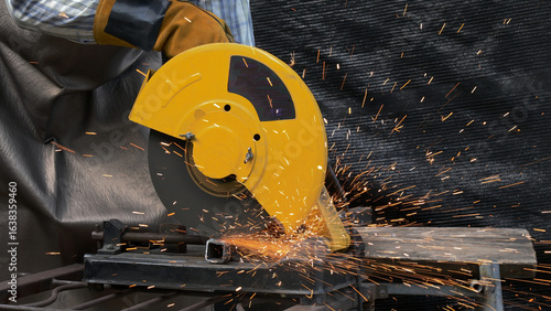 Close-up view of yellow chop saw cutting through square metal tube with bright sparks flying, operator's gloved hand visible