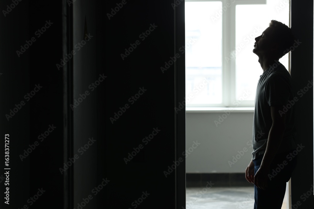 Fototapeta premium Sadness and depression. Tired man in dark room with open door against window with daylight. Space for text