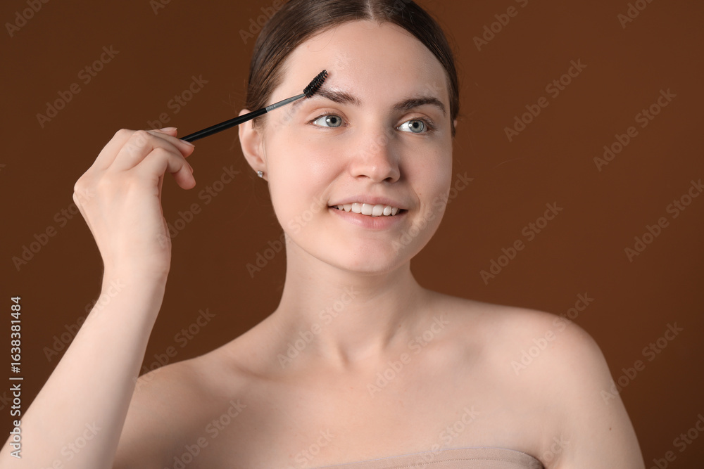 Young woman brushing her eyebrow with spoolie on brown background Young woman brushing her eyebrow with spoolie on brown background