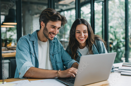 A happy male and female team member, sitting at an office desk with laptop open in front of them looking over the lap top screen smiling together while they work on their company website