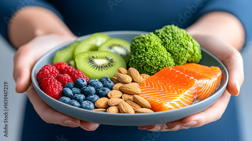 A healthy elimination diet plate filled with salmon, fruits, vegetables, and seeds is held indoors by a young woman.
