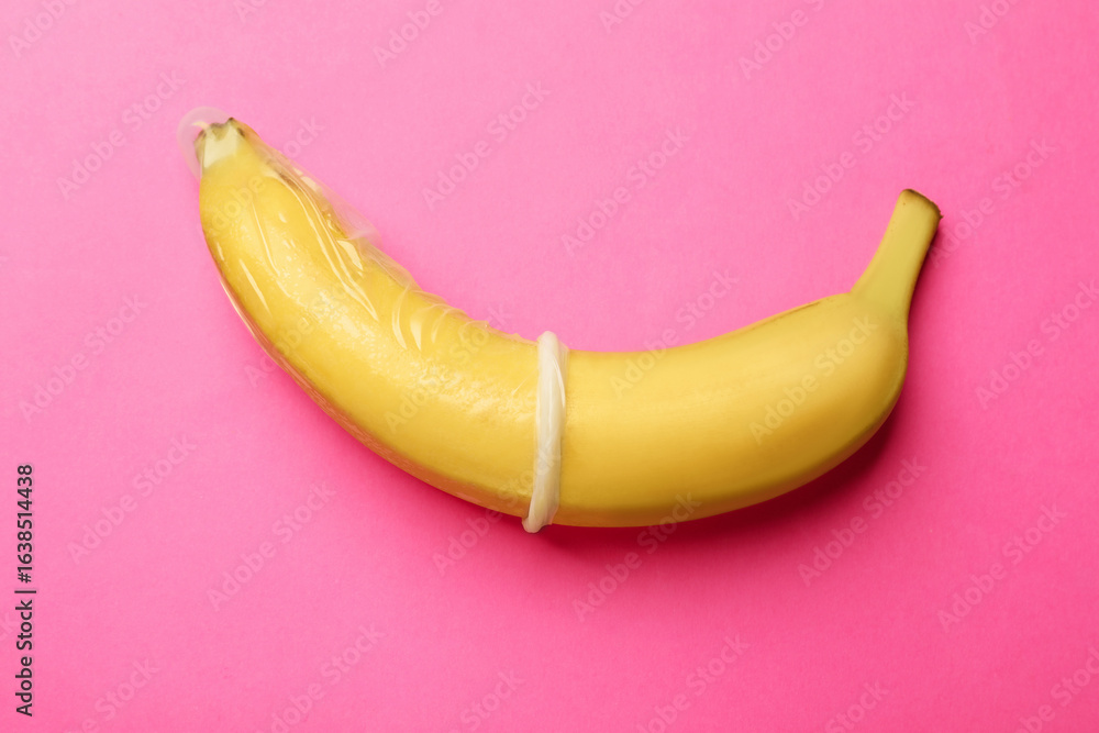 Fototapeta premium Sex education. Banana with condom on pink background, top view