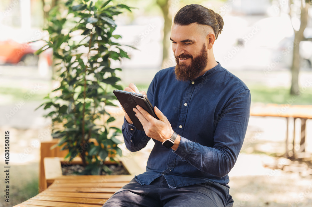 Obraz premium A man with a prominent beard is fully engaged with his tablet while enjoying a peaceful outdoor setting