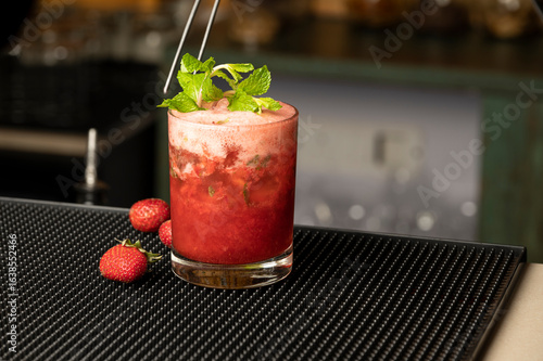 Close-up shot of strawberry-flavoured drink in a highball glass on bar mat, with fresh strawberries nearby and bartender placing mint garnish with tongs, blurred bar background