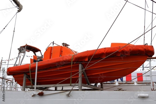 Fotografie An orange lifeboat hanging on the side of a tall ship