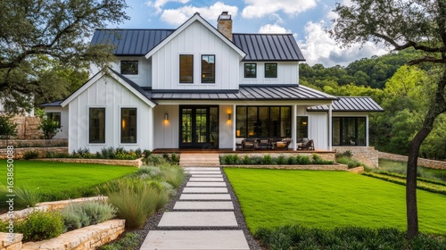 A white farmhouse with vertical siding, stone base, black metal roof, gabled forms, large windows, and vibrant green landscape.