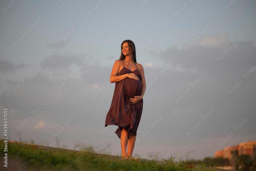 Obraz premium Smiling pregnant woman holding belly while standing on grassy slope under soft golden evening sunlight