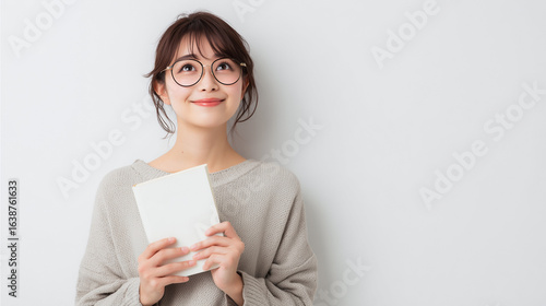 明るい白背景の前で、眼鏡をかけた若い日本人女性が表紙が白紙の白い本を両手で持ち、微笑みながら右上を見上げている。人物は画面の右下隅に配置され、左上には広々とした余白がある。シンプルで清潔感のあるポートレート。本を持つ, 白い壁, 読書, 考える