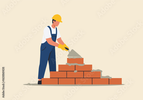 A professional builder in a hard hat and overalls constructing a red brick wall. The skilled builder uses a trowel to apply mortar.