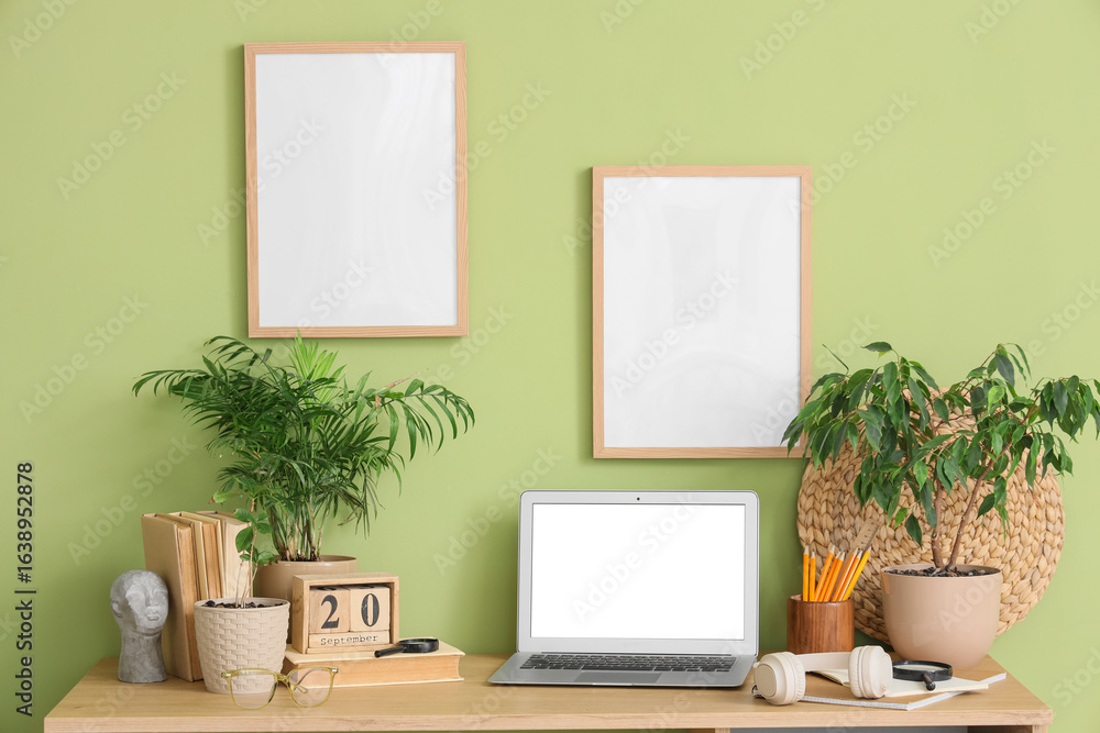 Obraz premium Comfortable workspace with modern laptop, headphones and houseplants near green wall