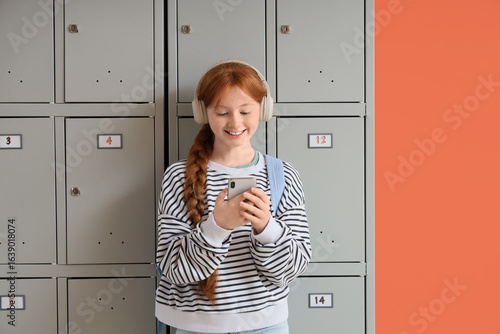 Teenage schoolgirl in headphones using mobile phone near locker on orange bac...