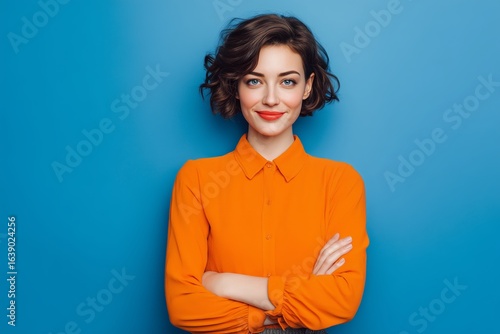 Attractive woman with bobbed hairstyle and crossed arms exuding confidence and friendliness in an orange office shirt against a blue background smile warm demeanor