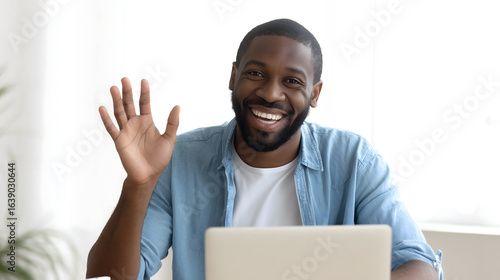 African American man is sitting at his desk, looking into the laptop screen and waving with one hand to say hi. His face shows happiness.