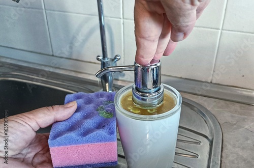 Pumping Dish Soap onto a Sponge. Hands and Kitchen Cleaning Supplies. ​Preparing to Wash Dishes. ​Close-up of a Soap Dispenser and Sponge.