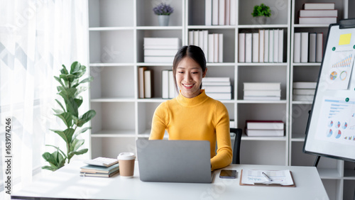 Young Asian Woman Working From Home on Laptop Computer Smiling and Confident. Home Office Setup with Bookshelf and Financial Report Board. Remote Work and Online Communication