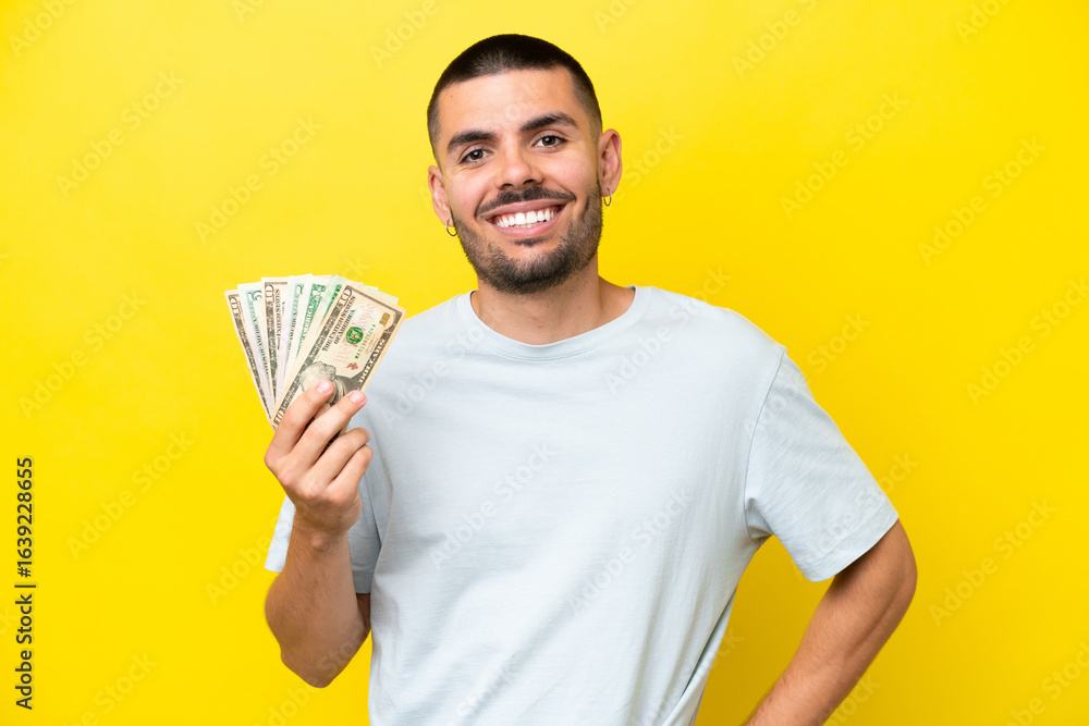 Young caucasian man taking a lot of money isolated on yellow background posing with arms at hip and smiling Young caucasian man taking a lot of money isolated on yellow background posing with arms at hip and smiling