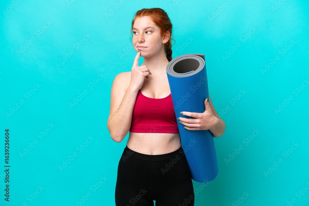 Young sport reddish woman going to yoga classes while holding a mat isolated on blue background having doubts while looking up Young sport reddish woman going to yoga classes while holding a mat isolated on blue background having doubts while looking up