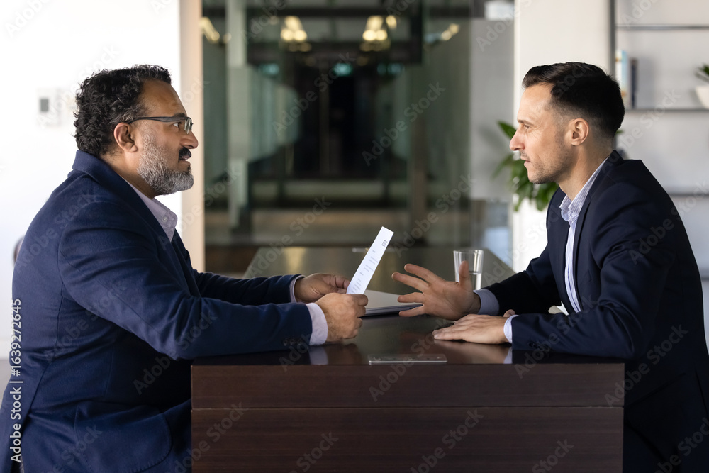 Fototapeta premium Two diverse male younger and older business coworkers arguing at work table, sitting opposite, talking, having conflict of generations. Senior boss dissatisfied with young professional paper report