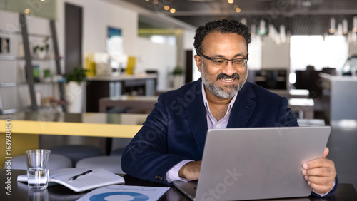 Foto Positive successful senior Indian business professional man typing on laptop, using computer for communication in co-working hall, working on Internet ebusiness project