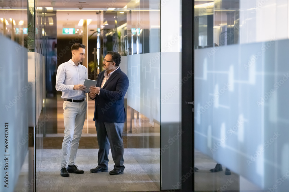 Fototapeta premium Elder Indian boss and younger manager discussing work on online project, using digital tablet together, standing in office corridor with glass walls, talking. Vertical full length shot
