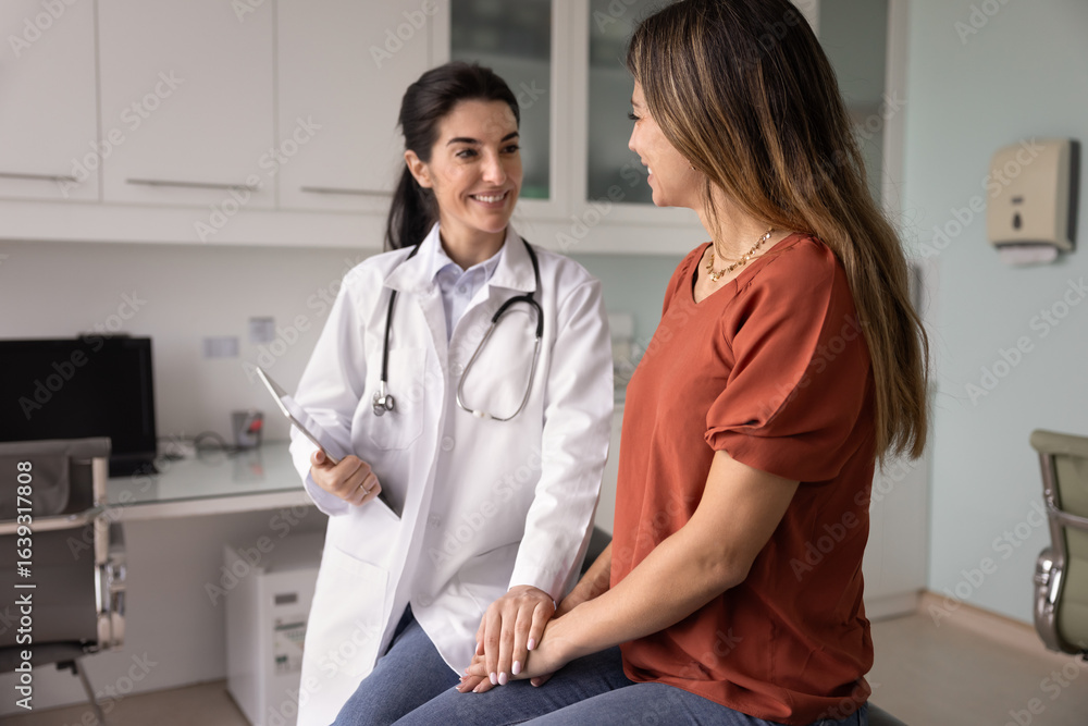 Obraz premium Happy optimistic young Latin physician meeting with patient after examination, touching hands of woman in supportive gesture, telling good news from medical report, smiling