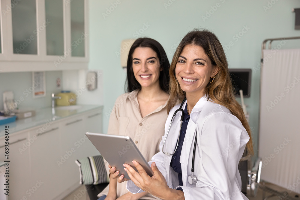 Happy successful young Latin doctor woman holding tablet, sitting on couch by positive female patient woman, looking at camera with toothy smile, posing for healthcare professional portrait Happy successful young Latin doctor woman holding tablet, sitting on couch by positive female patient woman, looking at camera with toothy smile, posing for healthcare professional portrait