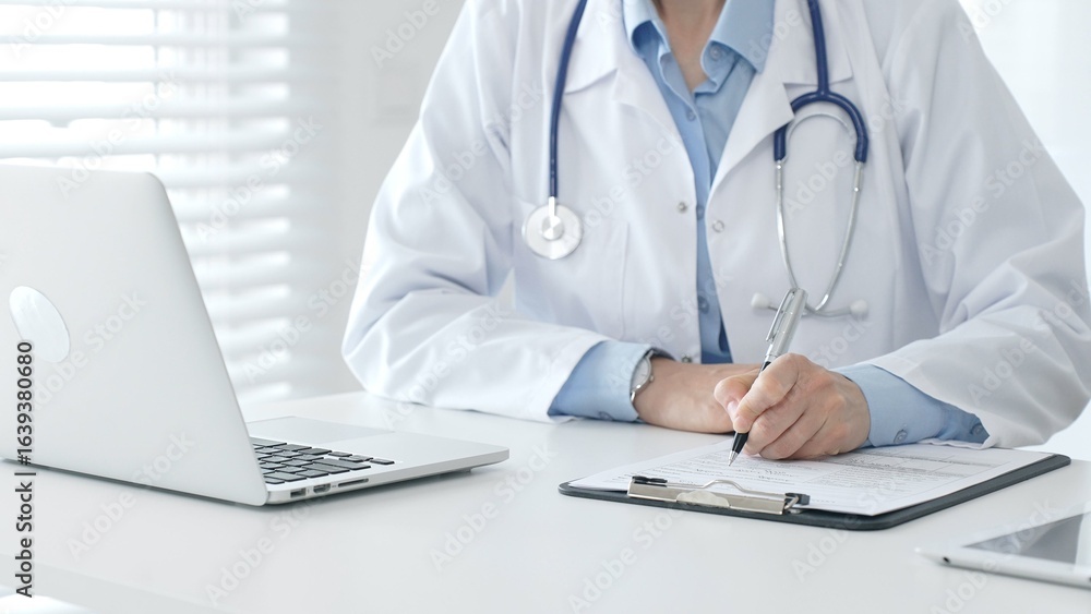 Female doctor documenting patient information while working with digital devices in a minimalist white workspace. Medicine and health care concept