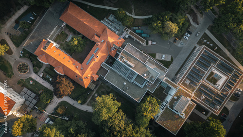 Aerial top-down view of a building complex with a red brick roof and modern structures with solar panels, surrounded by trees and green areas, captured in the evening light.
