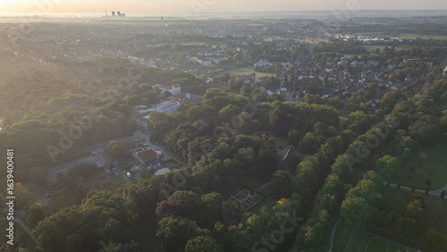 Aerial view of a city surrounded by dense green forest at sunrise, with soft morning light and mist in the distance, highlighting urban and natural landscapes.